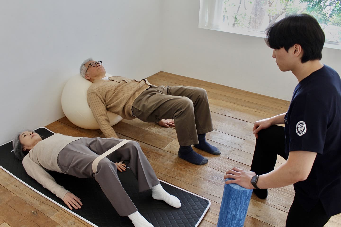 선생님이 짐볼과 밴드로 두 어르신의 브릿지 운동을 지도하는 방문재활 수업 장면