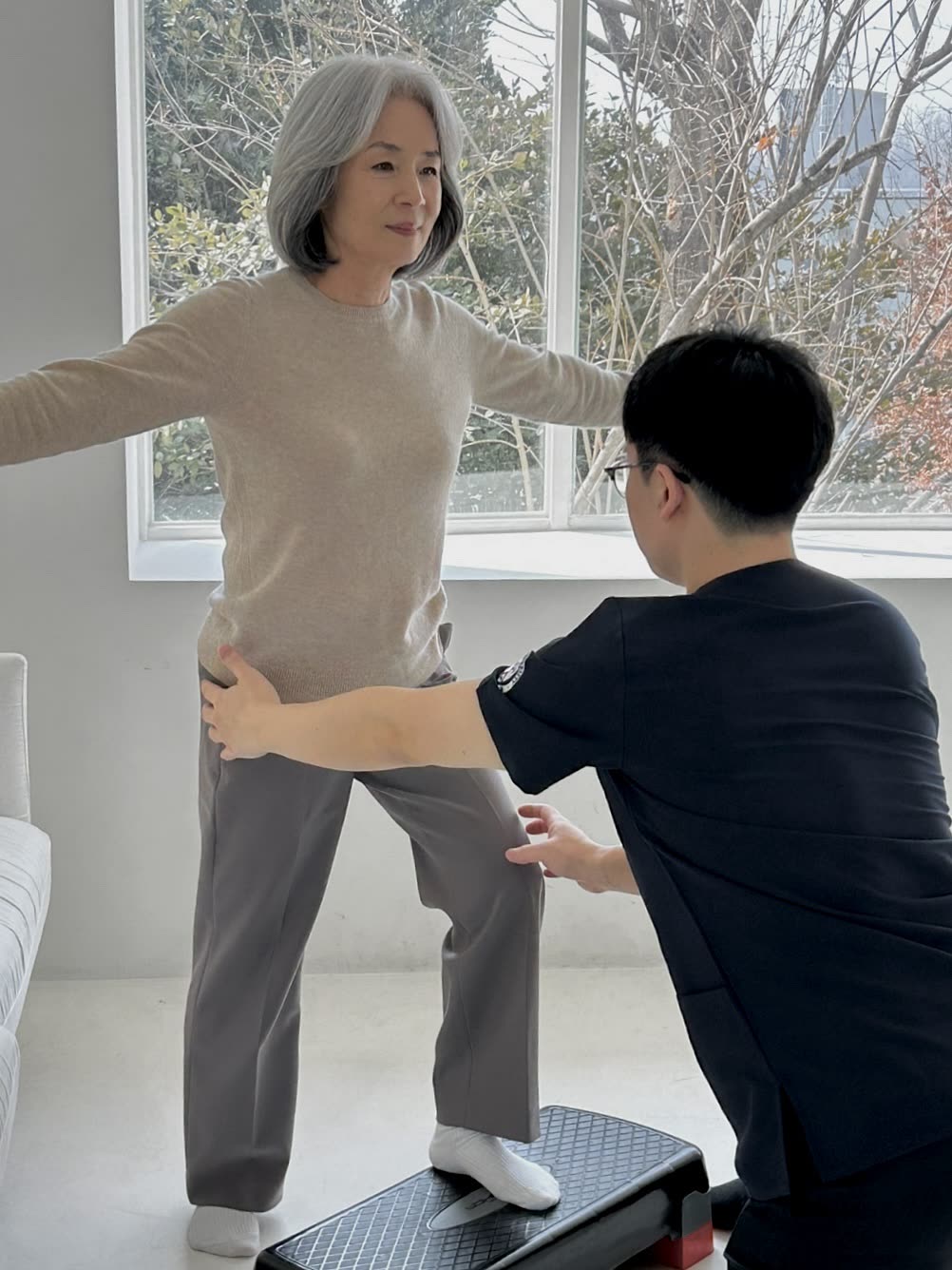 스텝 위에서 팔을 벌려 균형을 잡는 어르신을 선생님이 골반을 받쳐 돕는 장면